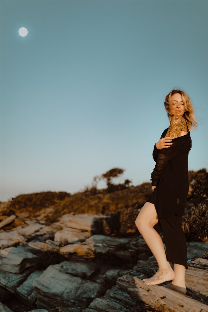 A woman on a windswept rocky Maine coast with the moon in the background