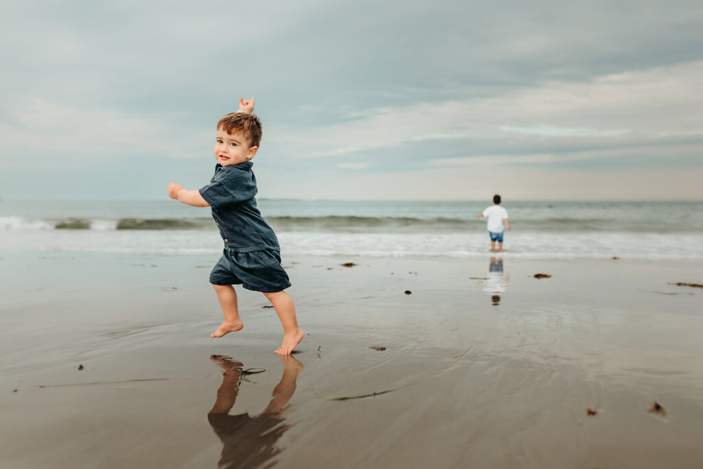 A young boy running on Old Orchard Beach in Maine while his brother looks at the ocean.