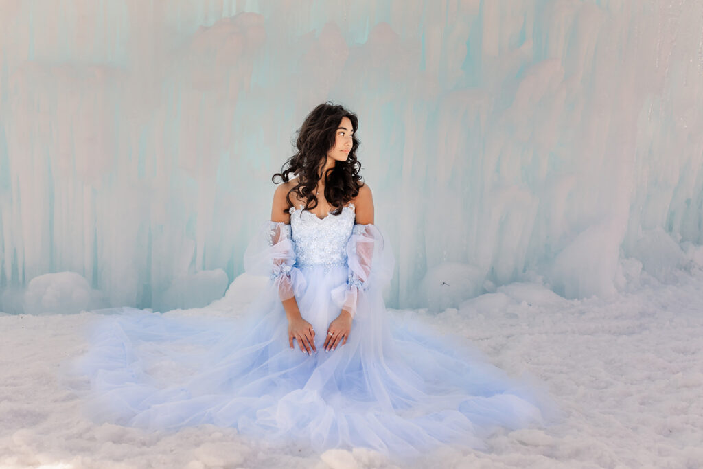 A girl kneeling in front of an ice wall at the New Hampshire Ice Castles in a blue, tulle gown