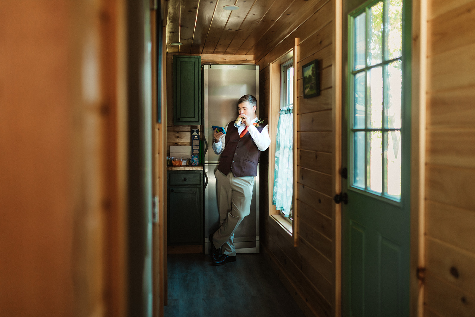 A groom casually leans against a door frame while avoiding seeing his bride and watching a soccer game while drinking a beverage