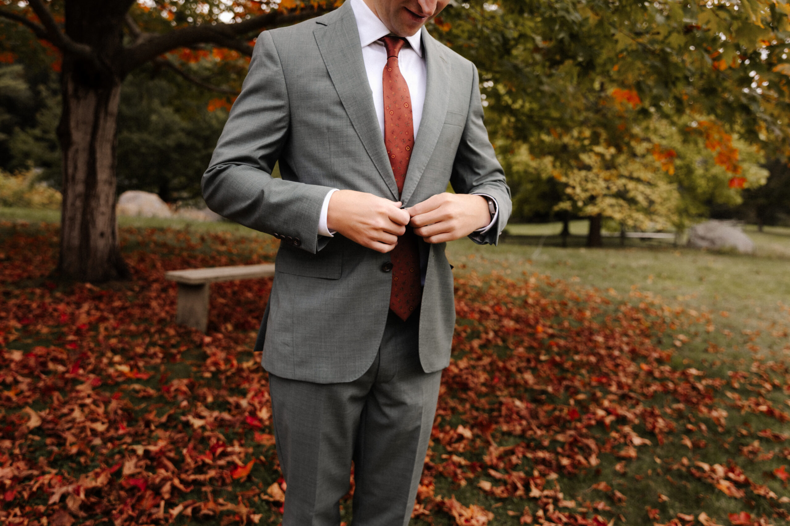 A groom buttoning his jacket against a colorful fall backdrop